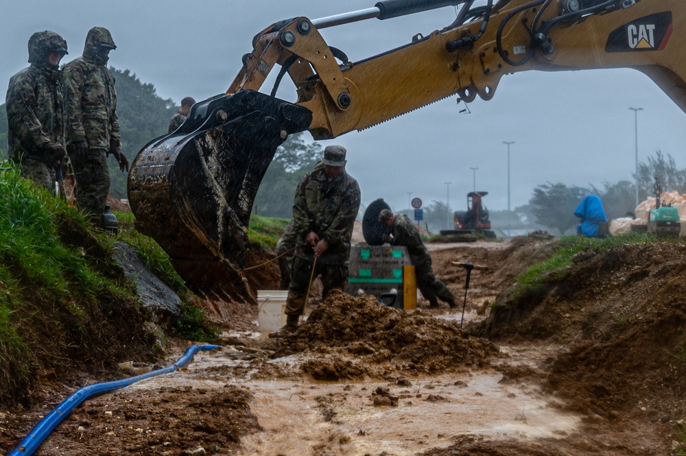 Military personnel in camouflage uniforms are conducting excavation and construction work in heavy rain using a CAT (Caterpillar) excavator. Workers are operating in muddy, waterlogged conditions, suggesting emergency infrastructure repair or construction operations. The scene depicts a coordinated effort with multiple personnel and heavy equipment working simultaneously in adverse weather.