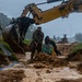 Military personnel in camouflage uniforms are conducting excavation and construction work in heavy rain using a CAT (Caterpillar) excavator. Workers are operating in muddy, waterlogged conditions, suggesting emergency infrastructure repair or construction operations. The scene depicts a coordinated effort with multiple personnel and heavy equipment working simultaneously in adverse weather.