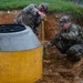 Two military personnel in OCP (Operational Camouflage Pattern) uniforms are measuring a large concrete cylindrical structure, likely a manhole or drainage unit, that has been partially installed in an excavated trench. One soldier leans over the top holding a tape measure while the other crouches at ground level holding the opposite end. The concrete structure has yellow and gray banding with Japanese text markings, suggesting this activity is taking place at a U.S. military installation in Japan.