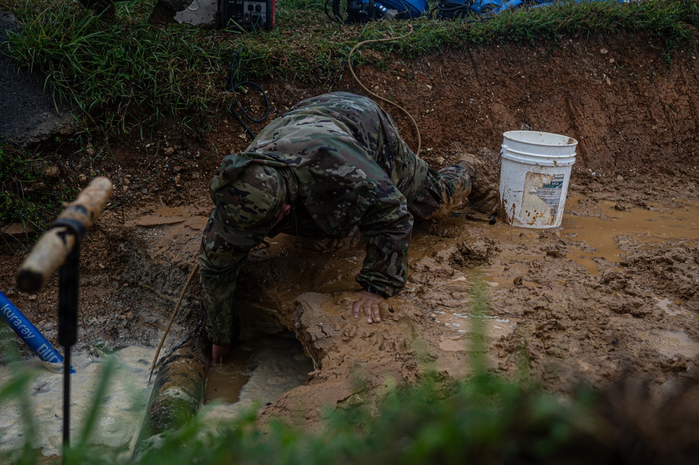 A person wearing military camouflage uniform is working in a muddy excavation pit, bent over and reaching down into standing water and mud. The scene appears to involve infrastructure repair work, likely on a buried pipe or utility line. The conditions are extremely muddy and wet, suggesting recent heavy rainfall or flooding.