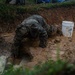 A person wearing military camouflage uniform is working in a muddy excavation pit, bent over and reaching down into standing water and mud. The scene appears to involve infrastructure repair work, likely on a buried pipe or utility line. The conditions are extremely muddy and wet, suggesting recent heavy rainfall or flooding.