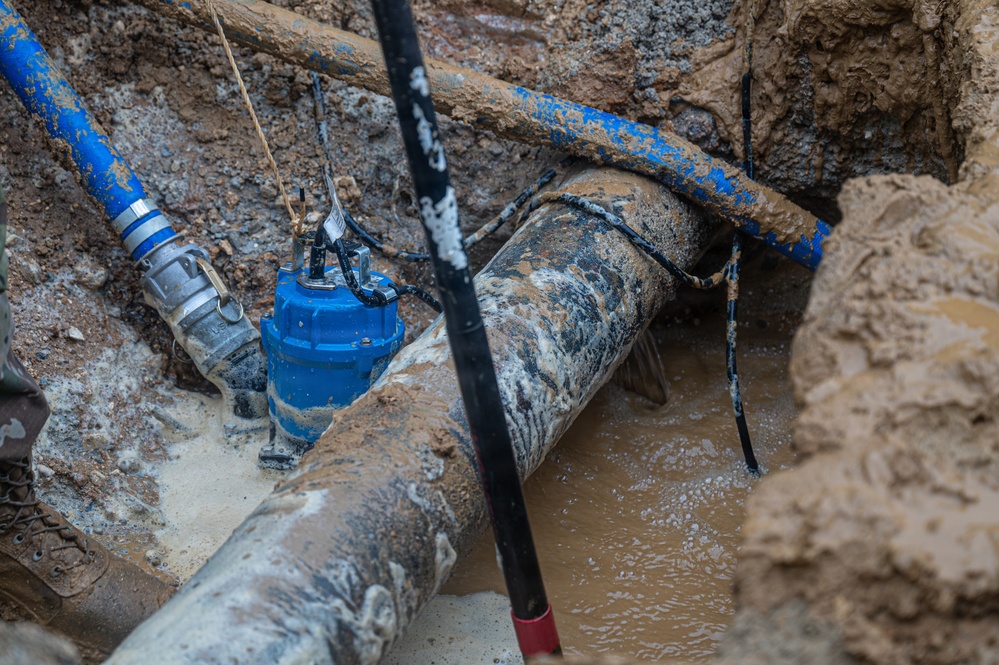The image shows an excavated trench containing underground utility infrastructure, including large corroded pipes and a blue submersible pump surrounded by muddy water. Workers appear to be conducting repair or replacement operations on what looks like aging water or sewer lines. The excavation reveals multiple pipe systems and temporary pumping equipment managing groundwater intrusion.
