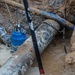 The image shows an excavated trench containing underground utility infrastructure, including large corroded pipes and a blue submersible pump surrounded by muddy water. Workers appear to be conducting repair or replacement operations on what looks like aging water or sewer lines. The excavation reveals multiple pipe systems and temporary pumping equipment managing groundwater intrusion.