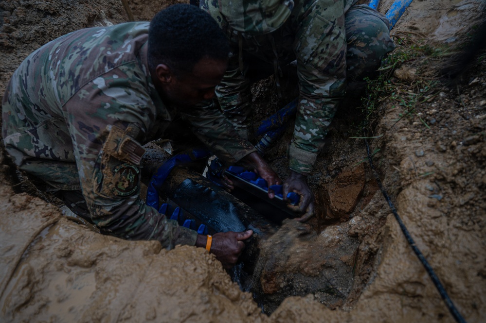 Two individuals wearing military camouflage uniforms are working in a dirt excavation trench, repairing or installing a water pipe. They appear to be applying a blue pipe repair clamp or coupling to a dark pipe buried underground. The scene suggests a military engineering or civil affairs water infrastructure repair operation.