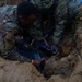 Two individuals wearing military camouflage uniforms are working in a dirt excavation trench, repairing or installing a water pipe. They appear to be applying a blue pipe repair clamp or coupling to a dark pipe buried underground. The scene suggests a military engineering or civil affairs water infrastructure repair operation.