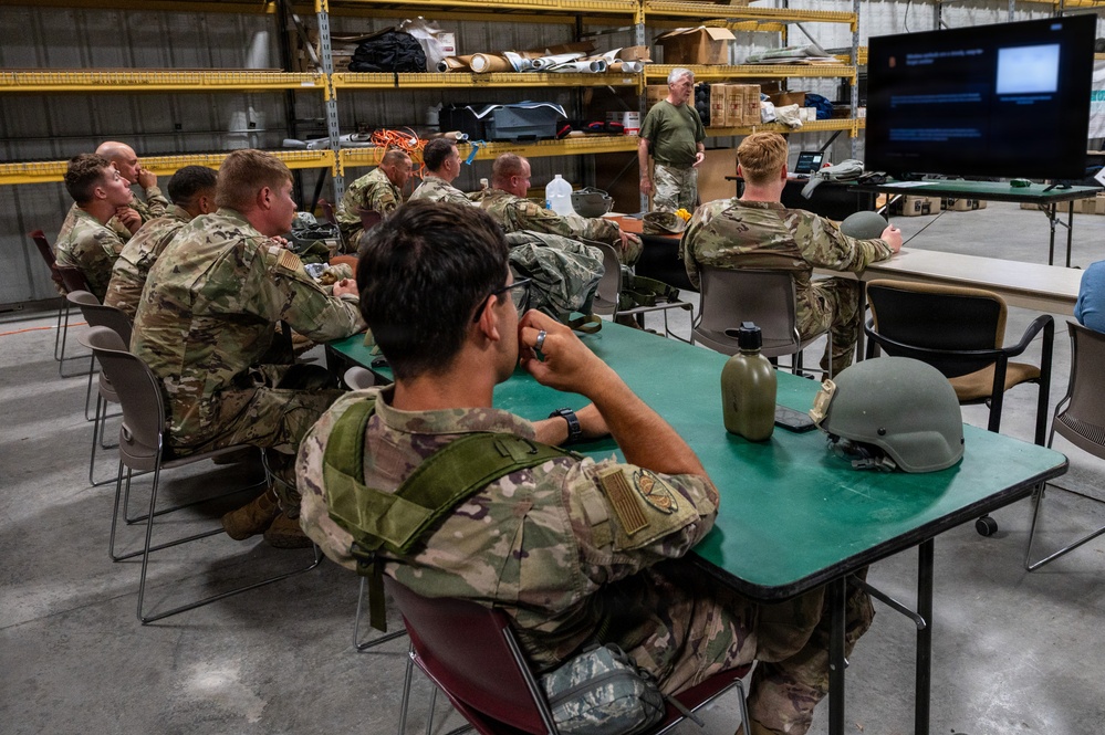A group of military personnel in camouflage uniforms are seated at tables in what appears to be a warehouse or hangar facility, attending a briefing or training session. An instructor stands at the front near a large display screen showing a presentation. Military equipment including helmets and canteens are visible on the tables.