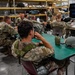 A group of military personnel in camouflage uniforms are seated at tables in what appears to be a warehouse or hangar facility, attending a briefing or training session. An instructor stands at the front near a large display screen showing a presentation. Military equipment including helmets and canteens are visible on the tables.