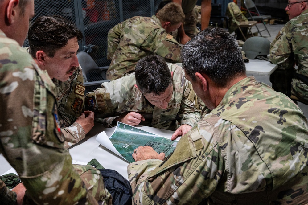 Several U.S. Air Force personnel in OCP (Operational Camouflage Pattern) uniforms are gathered around a table reviewing what appears to be an aerial photograph or map during a briefing or planning session. The central figures are leaning in closely to examine the document, suggesting mission planning or tactical coordination. Additional personnel are visible in the background engaged in similar activities.