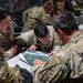 Several U.S. Air Force personnel in OCP (Operational Camouflage Pattern) uniforms are gathered around a table reviewing what appears to be an aerial photograph or map during a briefing or planning session. The central figures are leaning in closely to examine the document, suggesting mission planning or tactical coordination. Additional personnel are visible in the background engaged in similar activities.