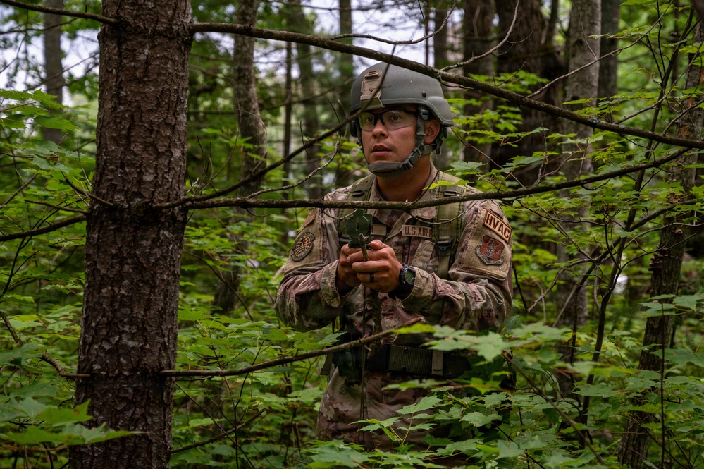 A U.S. Air Force service member in full combat gear navigates through a dense woodland area. The individual is wearing OCP (Operational Camouflage Pattern) uniform with visible patches including 'U.S. AIR' and 'HVAC' tape, a ballistic helmet, and protective eyewear. They appear to be holding a compass or small navigation device, suggesting land navigation training.