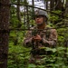 A U.S. Air Force service member in full combat gear navigates through a dense woodland area. The individual is wearing OCP (Operational Camouflage Pattern) uniform with visible patches including 'U.S. AIR' and 'HVAC' tape, a ballistic helmet, and protective eyewear. They appear to be holding a compass or small navigation device, suggesting land navigation training.