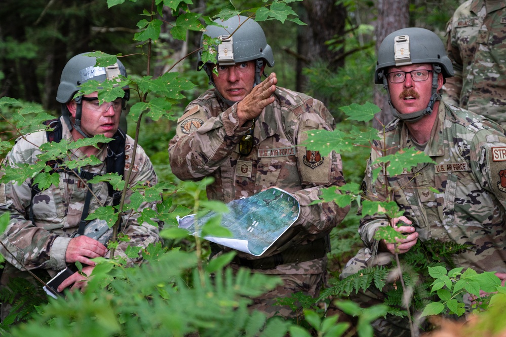 Three U.S. Air Force personnel in MultiCam combat uniforms and helmets are gathered in a wooded area, appearing to conduct a field briefing or mission planning session. The central figure is gesturing with his hand while holding what appears to be a printed aerial or satellite map. The setting suggests a field training exercise or operational planning scenario in a forested environment.