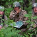 Three U.S. Air Force personnel in MultiCam combat uniforms and helmets are gathered in a wooded area, appearing to conduct a field briefing or mission planning session. The central figure is gesturing with his hand while holding what appears to be a printed aerial or satellite map. The setting suggests a field training exercise or operational planning scenario in a forested environment.