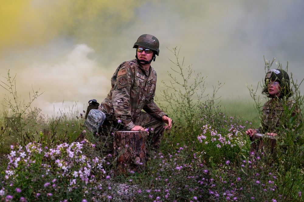 Two military personnel in camouflage uniforms and helmets are positioned in a field with wildflowers during what appears to be a training exercise. Colored smoke grenades have been deployed, creating yellow and white smoke clouds in the background. The soldiers are wearing protective eyewear and appear to be observing or assessing their surroundings.