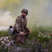 Two military personnel in camouflage uniforms and helmets are positioned in a field with wildflowers during what appears to be a training exercise. Colored smoke grenades have been deployed, creating yellow and white smoke clouds in the background. The soldiers are wearing protective eyewear and appear to be observing or assessing their surroundings.