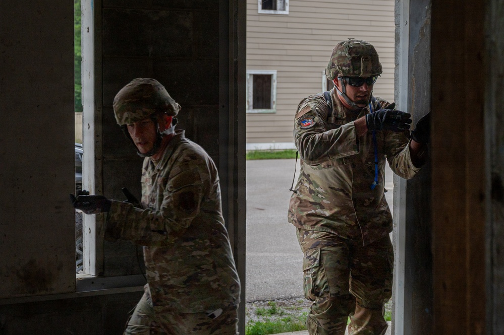 Two military personnel in OCP (Operational Camouflage Pattern) uniforms are conducting a building entry or clearing exercise at what appears to be a training facility. Both soldiers are wearing combat helmets and tactical gear, with one holding a weapon and moving through a doorway. The setting appears to be a mock urban environment used for military training.