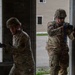 Two military personnel in OCP (Operational Camouflage Pattern) uniforms are conducting a building entry or clearing exercise at what appears to be a training facility. Both soldiers are wearing combat helmets and tactical gear, with one holding a weapon and moving through a doorway. The setting appears to be a mock urban environment used for military training.
