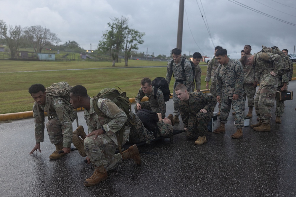 A group of military personnel in mixed camouflage uniforms (both US Army and US Marine Corps patterns) are conducting what appears to be a casualty evacuation or combat lifesaver training exercise on a wet road during overcast/rainy conditions. Several soldiers and Marines are crouched around a person lying on a litter/stretcher on the ground, while others stand nearby with rucksacks and gear. The setting appears to be a military installation.