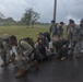 A group of military personnel in mixed camouflage uniforms (both US Army and US Marine Corps patterns) are conducting what appears to be a casualty evacuation or combat lifesaver training exercise on a wet road during overcast/rainy conditions. Several soldiers and Marines are crouched around a person lying on a litter/stretcher on the ground, while others stand nearby with rucksacks and gear. The setting appears to be a military installation.