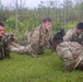 A group of military personnel in camouflage uniforms are performing low-crawl or push-up exercises on a grass field during what appears to be a training exercise. At least two distinct uniform patterns are visible — USMC MARPAT digital camouflage and US Army/Multicam OCP pattern — suggesting a joint or combined training event. The setting appears to be an outdoor training area enclosed by a wire fence, with overcast skies and lush vegetation in the background.