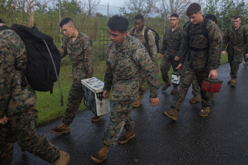 A group of military personnel in MARPAT camouflage uniforms are walking along a wet paved path, carrying various cases and equipment. The soldiers appear to be conducting a movement or logistics operation, possibly in a tropical environment given the vegetation visible in the background. The wet conditions suggest recent rainfall.