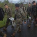A group of military personnel in MARPAT camouflage uniforms are walking along a wet paved path, carrying various cases and equipment. The soldiers appear to be conducting a movement or logistics operation, possibly in a tropical environment given the vegetation visible in the background. The wet conditions suggest recent rainfall.