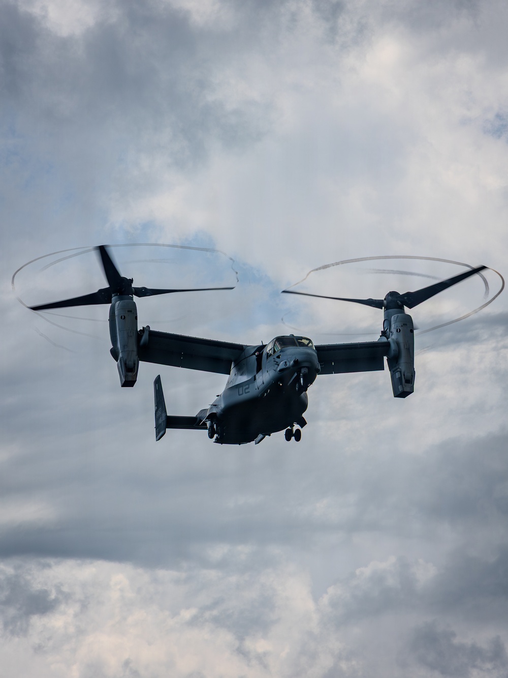 A military tiltrotor aircraft, identifiable as a Bell Boeing V-22 Osprey, is photographed from below against an overcast sky. The aircraft is in flight with both proprotors spinning at high speed, creating visible rotor blur circles. A tail number or side number '02' is visible on the fuselage.