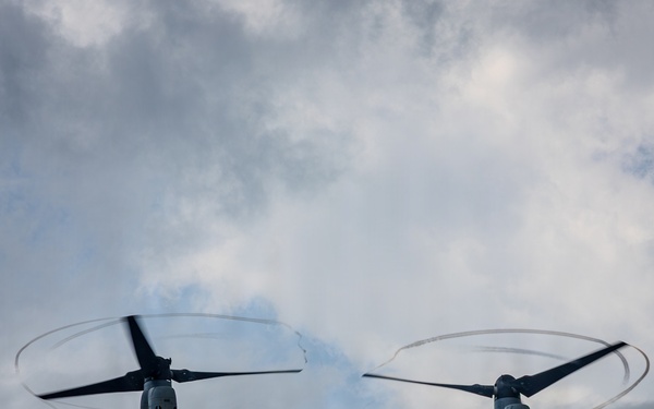 A military tiltrotor aircraft, identifiable as a Bell Boeing V-22 Osprey, is photographed from below against an overcast sky. The aircraft is in flight with both proprotors spinning at high speed, creating visible rotor blur circles. A tail number or side number '02' is visible on the fuselage.