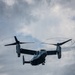 A military tiltrotor aircraft, identifiable as a Bell Boeing V-22 Osprey, is photographed from below against an overcast sky. The aircraft is in flight with both proprotors spinning at high speed, creating visible rotor blur circles. A tail number or side number '02' is visible on the fuselage.