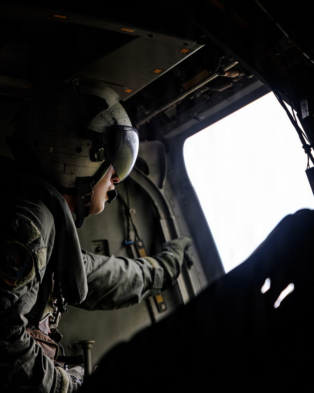 A military aircrew member wearing a flight helmet with visor is seated inside a helicopter, looking out through an open side window or door. The interior is dark and utilitarian, with the bright exterior sky creating strong contrast. The individual appears to be monitoring or observing the exterior environment during flight.