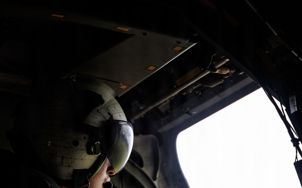 A military aircrew member wearing a flight helmet with visor is seated inside a helicopter, looking out through an open side window or door. The interior is dark and utilitarian, with the bright exterior sky creating strong contrast. The individual appears to be monitoring or observing the exterior environment during flight.