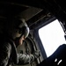 A military aircrew member wearing a flight helmet with visor is seated inside a helicopter, looking out through an open side window or door. The interior is dark and utilitarian, with the bright exterior sky creating strong contrast. The individual appears to be monitoring or observing the exterior environment during flight.