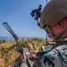 A military service member in MARPAT digital camouflage uniform is positioned in a concealed outdoor location among tall dry grass near a body of water. The individual is operating a handheld military radio (appears to be a Falcon III or similar tactical radio) while also referencing a ruggedized tablet device. The setting suggests a field training exercise or operational reconnaissance position.