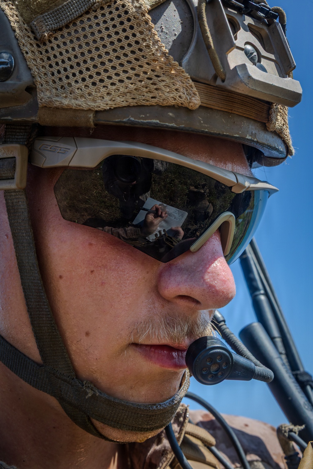 Close-up portrait of a military service member wearing tactical gear including a combat helmet, ESS brand ballistic eyewear, and a boom microphone. The sunglass lenses reflect another soldier appearing to review a document or map. The image is taken under bright daylight conditions with a clear blue sky visible in the background.