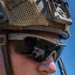 Close-up portrait of a military service member wearing tactical gear including a combat helmet, ESS brand ballistic eyewear, and a boom microphone. The sunglass lenses reflect another soldier appearing to review a document or map. The image is taken under bright daylight conditions with a clear blue sky visible in the background.