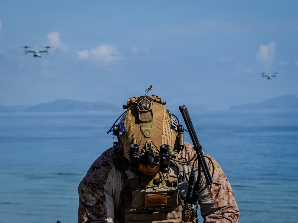A heavily equipped military operator in desert MARPAT camouflage is positioned on a beach, facing downward while operating communications or targeting equipment. Two V-22 Osprey tiltrotor aircraft are visible flying in the background over open water. The scene appears to depict an amphibious or littoral military exercise.