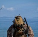 A heavily equipped military operator in desert MARPAT camouflage is positioned on a beach, facing downward while operating communications or targeting equipment. Two V-22 Osprey tiltrotor aircraft are visible flying in the background over open water. The scene appears to depict an amphibious or littoral military exercise.