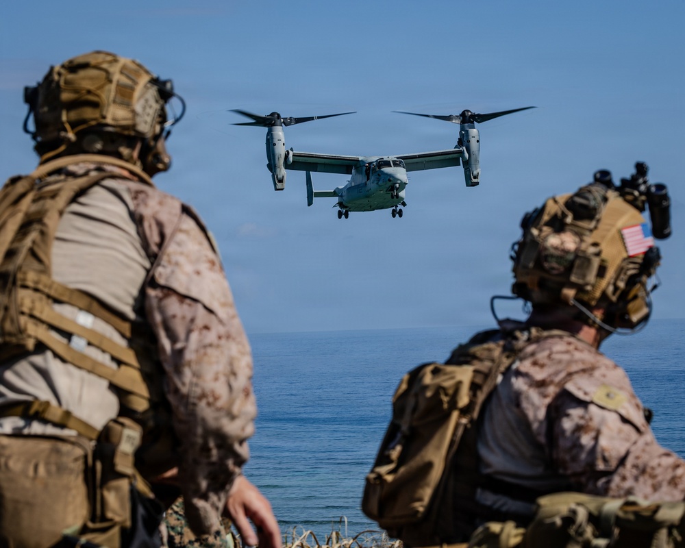 Two U.S. Marines in full combat gear observe a V-22 Osprey tiltrotor aircraft approaching over a body of water. The personnel are equipped with MARPAT desert camouflage uniforms, tactical helmets with accessories, and full combat load-bearing equipment. The scene appears to depict a military amphibious or coastal operation, likely an exercise or training evolution.