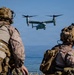 Two U.S. Marines in full combat gear observe a V-22 Osprey tiltrotor aircraft approaching over a body of water. The personnel are equipped with MARPAT desert camouflage uniforms, tactical helmets with accessories, and full combat load-bearing equipment. The scene appears to depict a military amphibious or coastal operation, likely an exercise or training evolution.