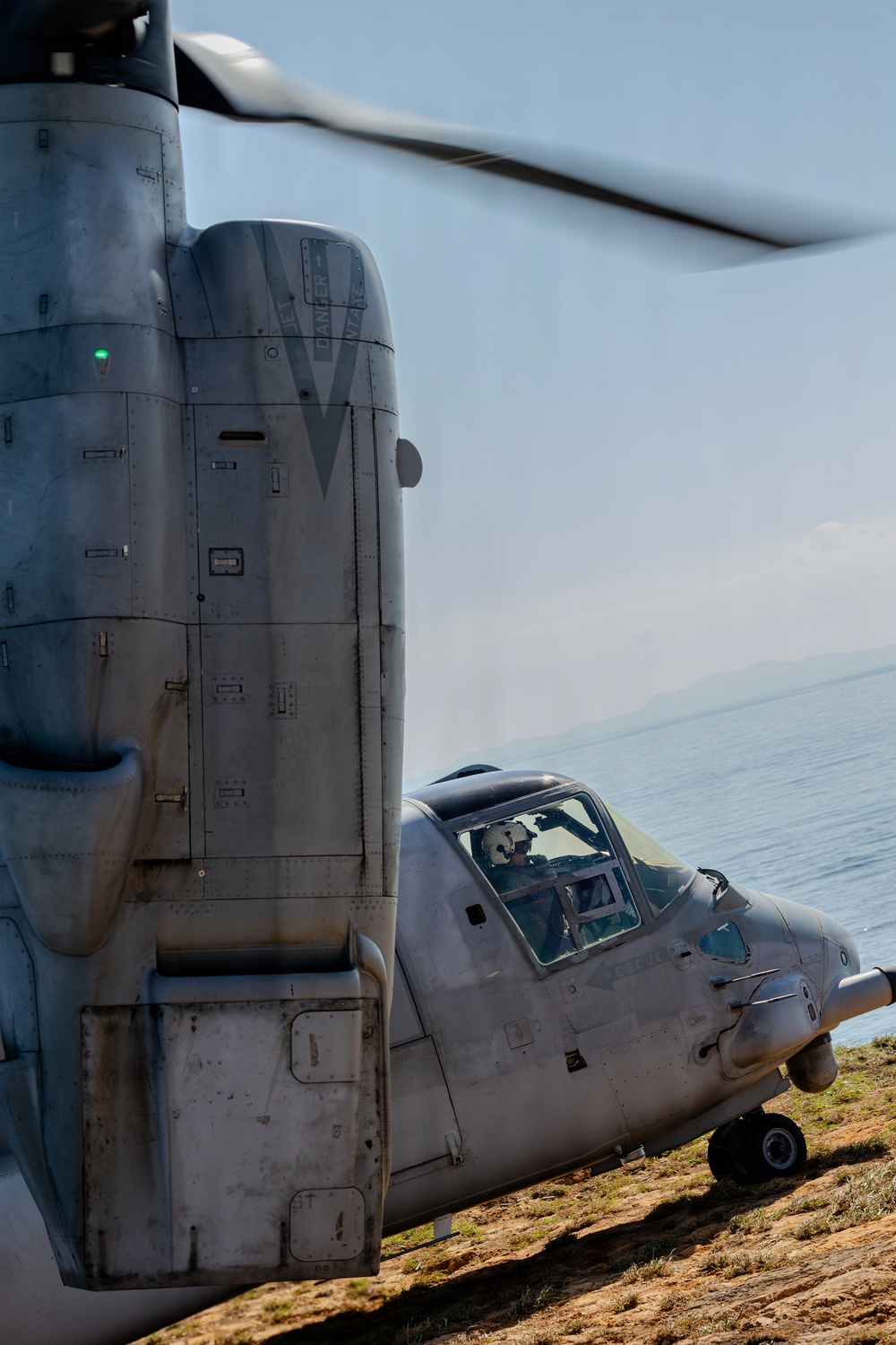 A large military tiltrotor aircraft, identifiable as a Bell Boeing V-22 Osprey, is shown on the ground near a coastal shoreline with rotors spinning. The aircraft is photographed from a close angle emphasizing the nacelle/engine housing and forward cockpit section. A helmeted crew member is visible through the cockpit windscreen.