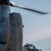 A large military tiltrotor aircraft, identifiable as a Bell Boeing V-22 Osprey, is shown on the ground near a coastal shoreline with rotors spinning. The aircraft is photographed from a close angle emphasizing the nacelle/engine housing and forward cockpit section. A helmeted crew member is visible through the cockpit windscreen.