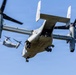 Two Bell Boeing V-22 Osprey tiltrotor aircraft are captured in flight against a clear blue sky. The foreground aircraft is prominently featured with its rear ramp open and a crew member visible in the doorway, landing gear deployed, suggesting a low-altitude operation. A second Osprey is visible in the background, slightly smaller, indicating formation flight.