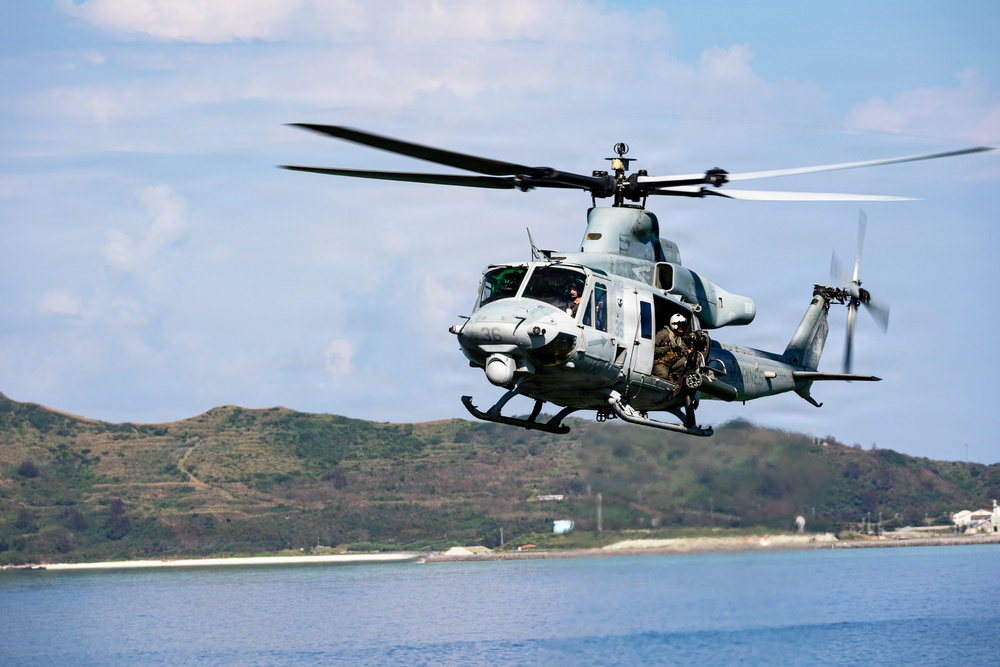 A military helicopter, identifiable as a UH-1Y Venom (or similar Bell UH-1 variant) in gray livery, is captured in low-level flight over a coastal body of water. At least two crew members are visible, with one appearing to be positioned at an open door. A green, hilly coastline and partial structures are visible in the background under a partly cloudy sky.