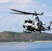 A military helicopter, identifiable as a UH-1Y Venom (or similar Bell UH-1 variant) in gray livery, is captured in low-level flight over a coastal body of water. At least two crew members are visible, with one appearing to be positioned at an open door. A green, hilly coastline and partial structures are visible in the background under a partly cloudy sky.