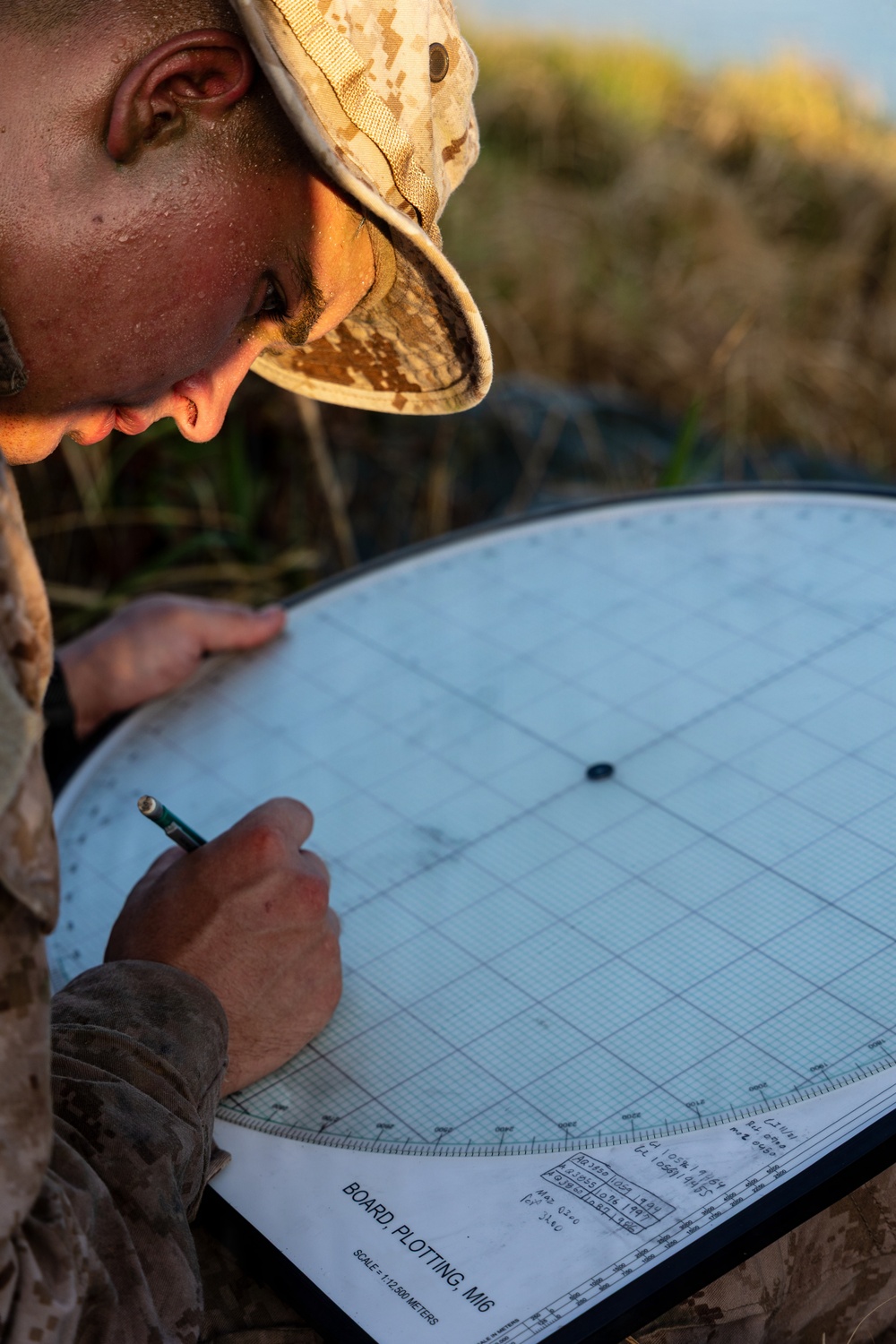 A military service member wearing desert MARPAT camouflage uniform and cover (boonie hat) is working with a M16 Plotting Board in an outdoor field environment. The individual is using a pencil to plot coordinates or bearings on the circular plotting board, which is labeled 'BOARD, PLOTTING, M16' with a scale of 1:12,500 meters. The setting appears to be during golden hour lighting with vegetation visible in the background.