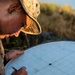 A military service member wearing desert MARPAT camouflage uniform and cover (boonie hat) is working with a M16 Plotting Board in an outdoor field environment. The individual is using a pencil to plot coordinates or bearings on the circular plotting board, which is labeled 'BOARD, PLOTTING, M16' with a scale of 1:12,500 meters. The setting appears to be during golden hour lighting with vegetation visible in the background.