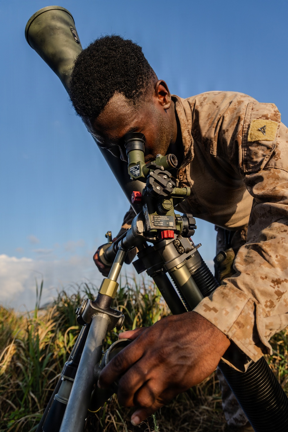 A military service member in desert MARPAT camouflage uniform is operating what appears to be an M64 mortar sight or similar optical aiming device mounted on a tripod. The individual is leaning in to look through the telescopic sight while adjusting the equipment with their hands. The setting appears to be an outdoor field environment with tall grass and a blue sky in the background.