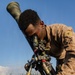 A military service member in desert MARPAT camouflage uniform is operating what appears to be an M64 mortar sight or similar optical aiming device mounted on a tripod. The individual is leaning in to look through the telescopic sight while adjusting the equipment with their hands. The setting appears to be an outdoor field environment with tall grass and a blue sky in the background.