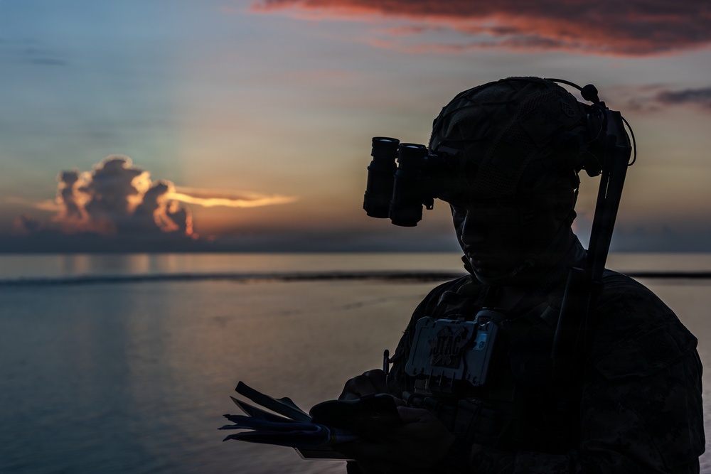 A silhouetted military personnel is photographed against a dramatic sunset over open water. The figure is equipped with tactical gear including a helmet-mounted night vision device and appears to be reviewing documents or a map. The low-light conditions create a high-contrast silhouette effect with vivid orange, pink, and blue tones in the background sky.