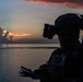 A silhouetted military personnel is photographed against a dramatic sunset over open water. The figure is equipped with tactical gear including a helmet-mounted night vision device and appears to be reviewing documents or a map. The low-light conditions create a high-contrast silhouette effect with vivid orange, pink, and blue tones in the background sky.
